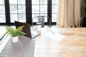 A wooden table with a laptop with a pile of paper, a pile and a glass of coffee on the background of a large window.
