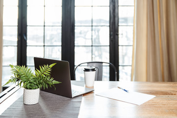 A wooden table with a laptop with a pile of paper, a pile and a glass of coffee on the background of a large window.