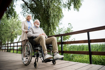 nice senior woman carrying husband in wheelchair while walking in park