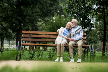 smiling senior couple embracing and holding hands while resting on wooden bench in park