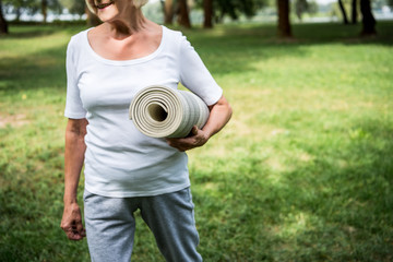 partial view of senior woman holding fitness mat while walking in park