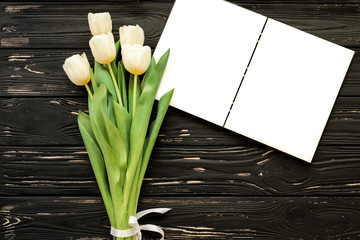 Beautiful bouquet of white tulips and open notebook on the black wooden background.
