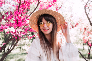 Happy pretty smiling young woman enjoying smell flowers,young beautiful woman near blooming tree, wearing straw hat and trendy shirt