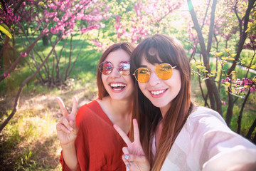 Image of young happy women friends standing in blossom spring park. Looking at camera make selfie.