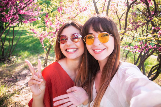 Image of young happy women friends standing in blossom spring park. Looking at camera make selfie.