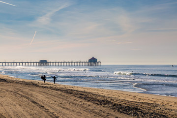 Silhouetted surfers walking along Huntington Beach in California, with the pier in the distance