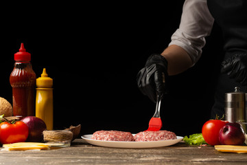 The cook rubs beef burgers with butter, against the background with ingredients, a restaurant business, fast food, tasty food, gastronomy, cooking, menu. Horizontal photo