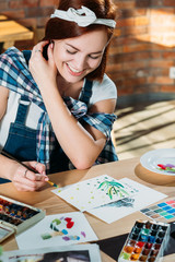 Fototapeta premium Studio workplace. Painting hobby. Smiling redhead woman doing first brushstrokes with palette supplies around.