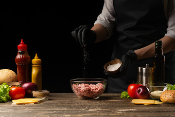 Chef salting mincemeat to create patties for a burger. Against the background with ingredients for a burger. The concept of facet food, tasty and fatty food. Gastronomy, recipes, menus