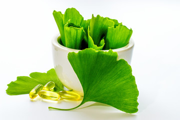 Healing leaves of ginkgo biloba tree in a white ceramic bowl and yellow capsules. Green leaves on a white background.	