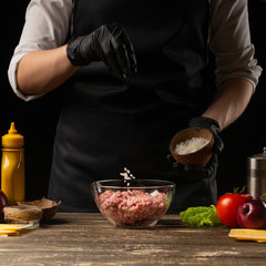 Chef salting minced meat to create a burger patty. Against the background with ingredients for a burger. Gastronomy, recipes, menus, fast food. Juicy hamburger