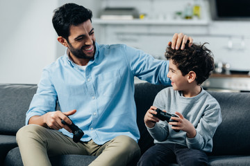 happy latin father touching hair of cute son and holding joysticks at home