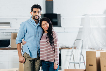 cheerful latin couple embracing and smiling near boxes in new home