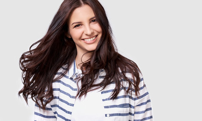 Fototapeta premium Closeup indoor portrait of beautiful brunette young woman with long hair smiling cheerfully. Charming female smile broadly showing her white healthy teeth to camera while feeling happy and carefree