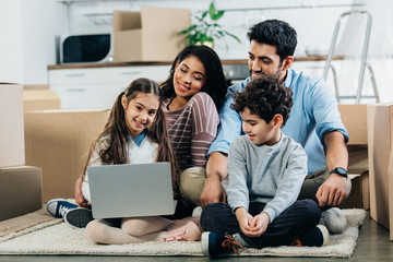 cheerful hispanic family looking at laptop while sitting on carpet in new home