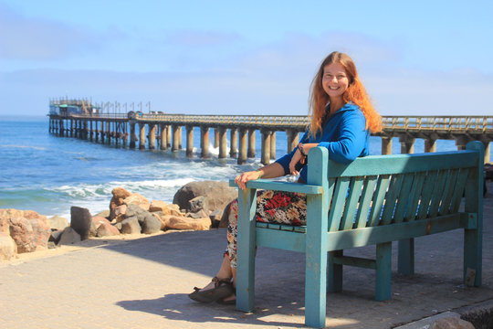 Red-haired happy white Caucasian girl sits on a bench on the shore of the Atlantic Ocean in Swakopmund (Namibia) and laughs on the background of the pier