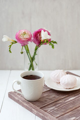 Spring, summer still life scene. A cup of tea in a white cup. Next is a bouquet with white freesia and pink ronunculuses. Next there is a white plate with pink marshmallow.