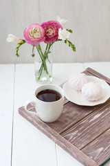 Spring, summer still life scene. A cup of tea in a white cup. Next is a bouquet with white freesia and pink ronunculuses. Next there is a white plate with pink marshmallow.