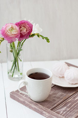 Spring, summer still life scene. A cup of tea in a white cup. Next is a bouquet with white freesia and pink ronunculuses. Next there is a white plate with pink marshmallow.