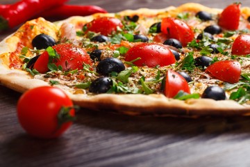 Close up Italian pizza with melted cheese, red cherry tomatoes, black olives, ham, and green leaves on a brown table decorated with mushrooms, red hot chili pepper and cherry tomatoes