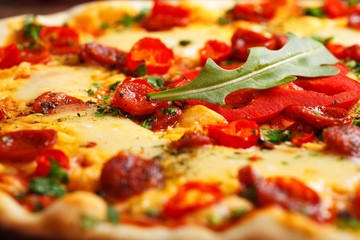 Close up Italian pizza with melted cheese, sausage, snack sticks, pepper and fresh green oregano leaves on a brown table decorated by mushrooms, red sweet pepper and cherry tomatoes