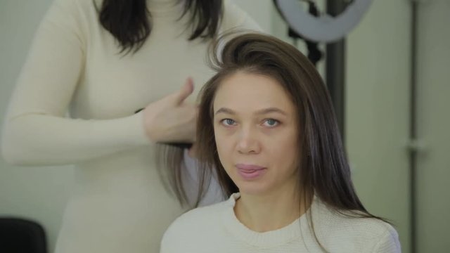 Beautiful woman with brown hair is having it treated with a curling iron by a hairdresser. Handheld real time establishing shot.