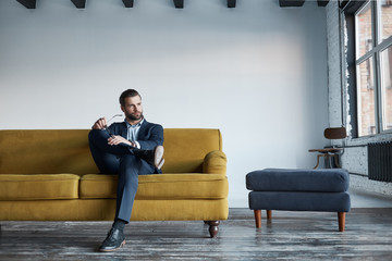 Portrait of bearded and handsome businessman in fashion suit who is resting on sofa in a modern...