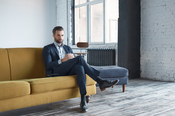 Business look: successful and handsome business man is sitting on office sofa and looking aside...