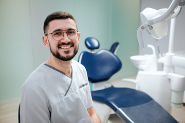 Obraz premium Positive young bearded dentist sit in dentistry room and pose on camera. He wear white robe and glasses.