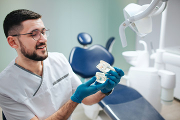 Cheerful young dentist sit in dentistry room. He hold and look at artificial jaw with teeth. Doctor wear glasses and smile.