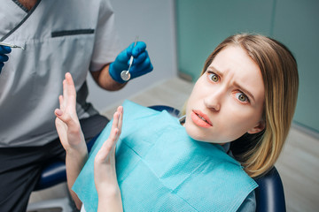 Young woman look afraid. She sit in chair in dentistry. Woman look on camera and push away doctor....