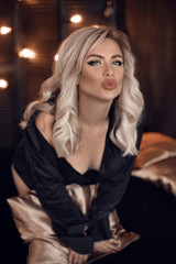 Fototapeta na wymiar Sexy blonde woman portrait in black shirt and lingerie. Beautiful fashion blond girl model over bokeh lights dark background. Alluring female with makeup and curly hair style posing on bed.