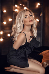 Naklejka na ściany i meble Sexy blonde woman portrait in black shirt and lingerie. Beautiful fashion blond girl model over bokeh lights dark background. Alluring female with makeup and curly hair style posing on bed.