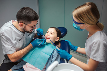 Male dentist check up girl's teeth with dentist's tools. Female helper stand beside. Girl sit in dental chair in room. She keep mouth opened.