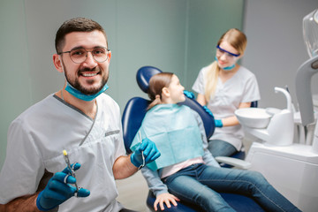 Obraz premium Happy male dentist look on camera and smile. He hold tools. Female dentist stand at girl. Child sit in dental chair.