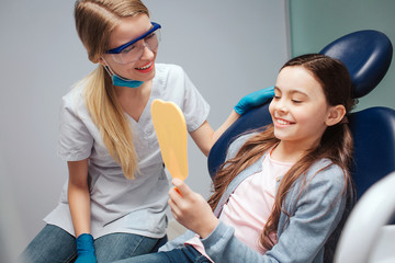 Hapy girl sit in dental chair in room. She look in mirror and smile. Female dentist sit beside her....