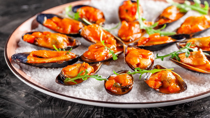 European cuisine. Marinated mussels in tomato sauce with rosemary, garlic, chili. Serving dishes in the restaurant on a plate with coarse sea salt on a black background. copy space