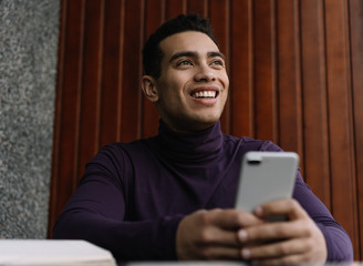 Happy handsome African American man holding mobile phone using application for online shopping with low prices, discount sales, cash back. Positive millennial guy waiting for taxi in modern cafe. 