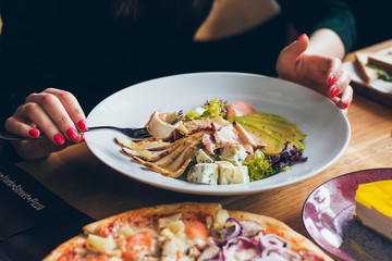 A woman eating tasty fresh salad with different ingredients like salad, tomatoes, meat, cheese in the restaurant with fork