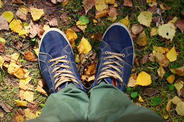 Teenage legs in sneakers and jeans standing on ground with autumn leaves, top view.