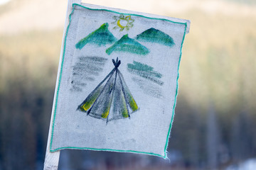 Camping flag showing a simple indian tent with mountains and sun in the back, hand drawn on a white hemp cloth. Blurry background with grass, during Spring season. Outdoor music festival concept.