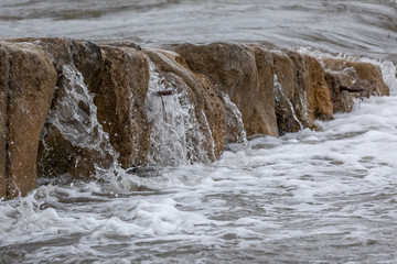 Splashing wave on rock wall in sea with metal mooring eye