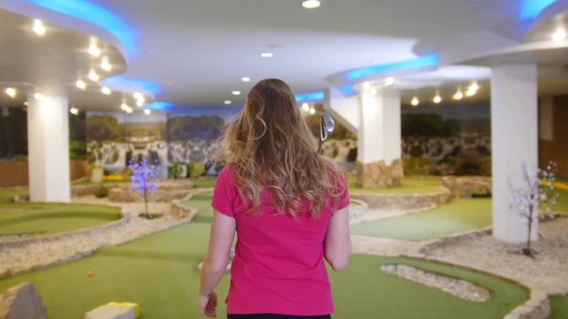 Playing mini golf. A young blonde woman takes a golf stick from the bag and goes away