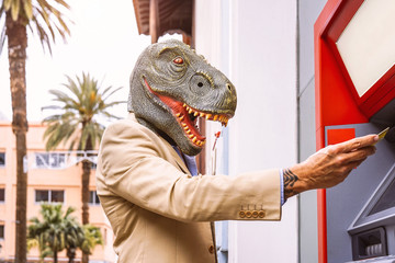 Senior man wearing t-rex dinosaur mask withdraw money from bank cash machine with debit card -...