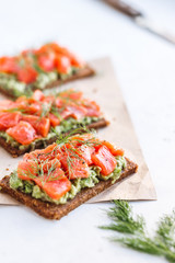 Close-up view of three sandwiches with rye bread, avocado and smoked salmon on a white kitchen table.