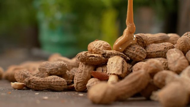 SLOW MOTION, MACRO, DOF: Creamy peanut butter is flowing onto the dry peanut shells scattered on the table. Mouth-watering shot of delicious nut butter creating a swirl on top of dry peanut shells.
