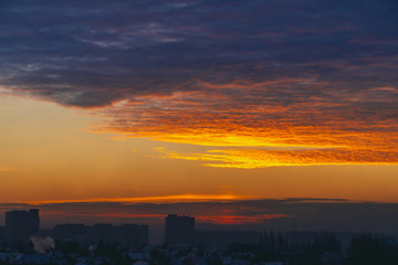 Cityscape with wonderful varicolored fiery dawn. Amazing dramatic multicolored cloudy sky. Dark silhouettes of city building roofs. Atmospheric background of sunrise in overcast weather. Copy space.