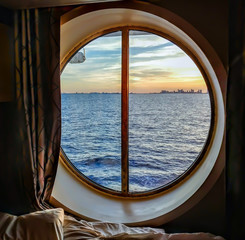 A view from the porthole window of a cruise ship, showing the city buildings in the sunset. The photo was taken inside a cabin on a Caribbean cruise.