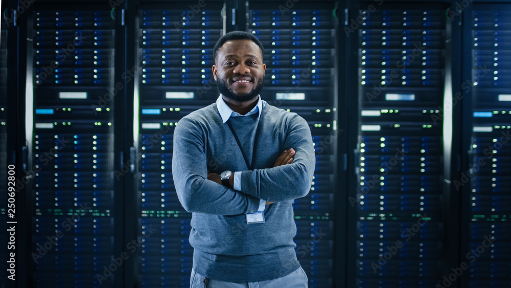 Poster Bearded Black IT Engineer Standing and Posing with Crossed Arms in the Middle of a Working Data Center Server Room with Server Computers Working on a Rack. 