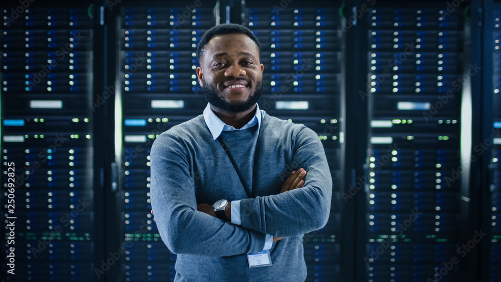 Poster Bearded Black IT Engineer Standing and Posing with Crossed Arms in the Middle of a Working Data Center Server Room with Server Computers Working on a Rack. 